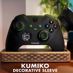 May include: Black Xbox controller with green accents, resting on a decorative wooden stand. The controller features colored buttons and the Foxwood logo. The stand has a geometric pattern and the text "KUMIKO DECORATIVE SLEEVE".