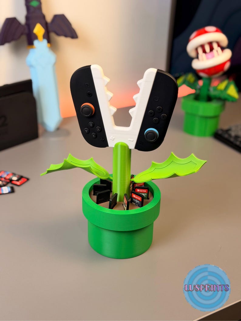 May include: A green, 3D-printed planter shaped like a pipe, holding Nintendo Switch game cartridges. The planter supports a stylized controller design with black and white Joy-Cons. A red and white Piranha Plant is in the background.