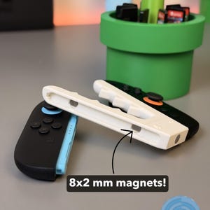 May include: A black and blue Nintendo Switch Joy-Con controller next to a white controller grip. The image also shows a green Mario-themed planter with game cartridges. The text in the image says "8x2 mm magnets!"