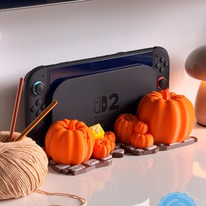 May include: A black Nintendo Switch console is displayed with several orange pumpkin decorations. A ball of yarn with knitting needles sits to the left. The pumpkins are of varying sizes and sit on a brown base.