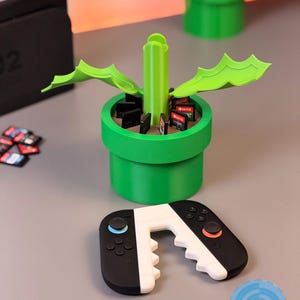 May include: A green, 3D-printed video game cartridge holder shaped like a pipe, with a matching controller. The holder contains game cartridges. The controller is black and white with blue and orange accents. The logo "LUSPRINTS" is visible.