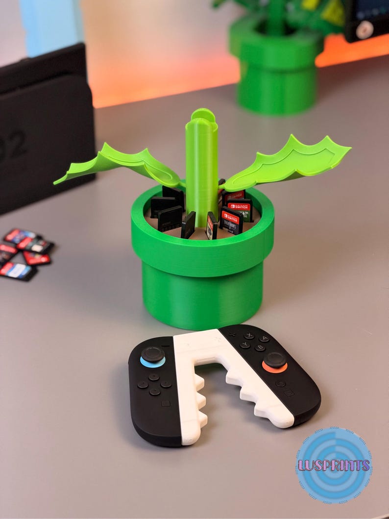 May include: A green, 3D-printed video game cartridge holder shaped like a pipe, with a matching controller. The holder contains game cartridges. The controller is black and white with blue and orange accents. The logo "LUSPRINTS" is visible.
