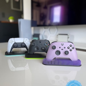 May include: Three video game controllers on stands. The controllers are white, black, and purple. The stands are green, yellow, and purple.