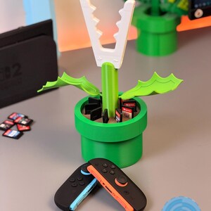 May include: A green and white 3D-printed Piranha Plant from the Super Mario video game series. The plant is in a green pipe, with a white mouth and green leaves. Two black and colored Switch controllers are in the foreground.