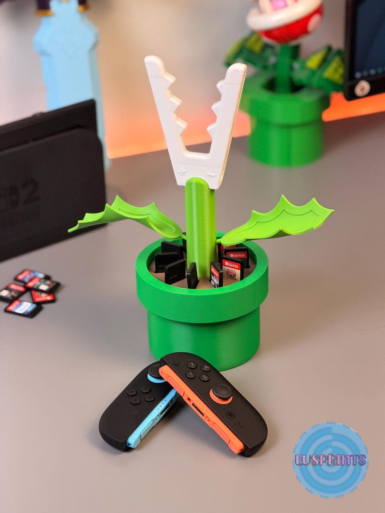 May include: A green and white 3D-printed Piranha Plant from the Super Mario video game series. The plant is in a green pipe, with a white mouth and green leaves. Two black and colored Switch controllers are in the foreground.