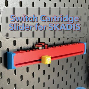 May include: A red and blue Switch cartridge slider for SKADIS, holding several game cartridges. The slider has a yellow question mark block attached. The text "Switch Cartridge Slider for SKADIS" is displayed on a clear background.