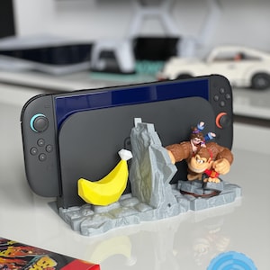 May include: A black Nintendo Switch console is docked in a custom stand featuring a yellow banana and a Donkey Kong-themed figurine. The stand is gray and has a rocky texture. A red game case is in the foreground.