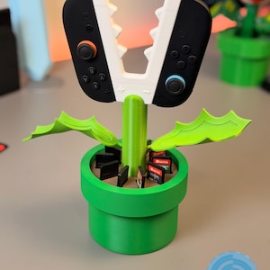 May include: A green, 3D-printed planter shaped like a Super Mario Bros. pipe, holding a controller-shaped game cartridge holder. The holder has a white and black design, resembling a Piranha Plant. Game cartridges are visible inside the planter.