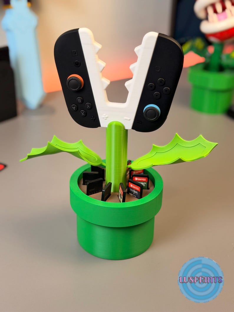 May include: A green, 3D-printed planter shaped like a Super Mario Bros. pipe, holding a controller-shaped game cartridge holder. The holder has a white and black design, resembling a Piranha Plant. Game cartridges are visible inside the planter.