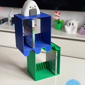 May include: A blue and green 3D printed video game case holder with a white onigiri figure on top. The holder has slots for video game cases and is designed to hold multiple games.