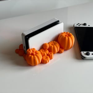 May include: A white Nintendo Switch dock with a cluster of orange pumpkin decorations. A Nintendo Switch console is also present, featuring a black screen and white controllers. The pumpkins vary in size and are clustered together.