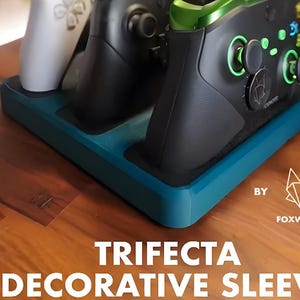 May include: A teal Trifecta decorative sleeve by Foxwood, holding three game controllers. The controllers are black, white, and green. The sleeve sits on a wooden surface.