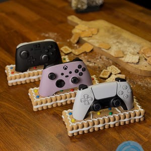 May include: Three game controllers in black, lavender, and white, each resting on a gingerbread-style base with colorful candy decorations. The controllers are on a wooden surface, with a cutting board and cookies in the background.