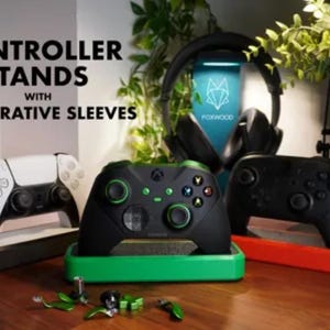 May include: Image of controller stands with decorative sleeves. The image shows three game controllers on stands, with a headset in the background. The controllers are black and white, black and green, and all black. The stands are black, green, and red.