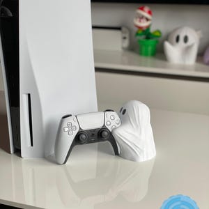 May include: A white PlayStation 5 console and controller are displayed next to a white ghost-shaped figurine. The controller has black accents. A small, green potted plant with a red and white flower is visible in the background.