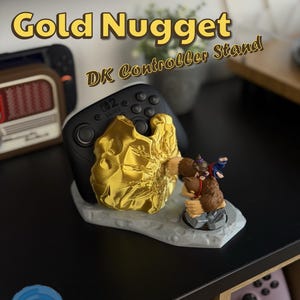 May include: A black Nintendo Switch controller rests on a gold-colored nugget-shaped stand. The stand features a Donkey Kong figurine. The text "Gold Nugget DK Controller Stand" is displayed in yellow.