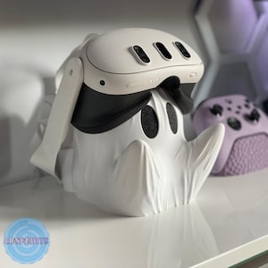 May include: A white VR headset rests on a white ghost-shaped stand. The headset has a black strap and three black lenses. A purple game controller is visible in the background. The stand has two black eye-shaped cutouts.
