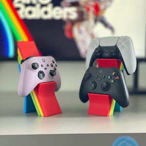 May include: Three game controllers, including a light purple Xbox controller, a black Xbox controller, and a white and black PlayStation controller, are displayed on colorful stands. The stands are red, yellow, blue, and green. The background shows the text "ARC Raiders".