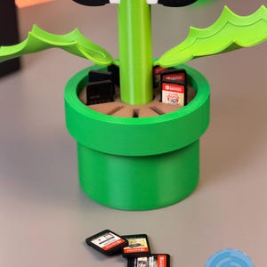 May include: A green and brown 3D-printed video game cartridge holder shaped like a flower pot, with a Nintendo Switch controller on top. Several game cartridges are in the pot, and three are in front of the pot.