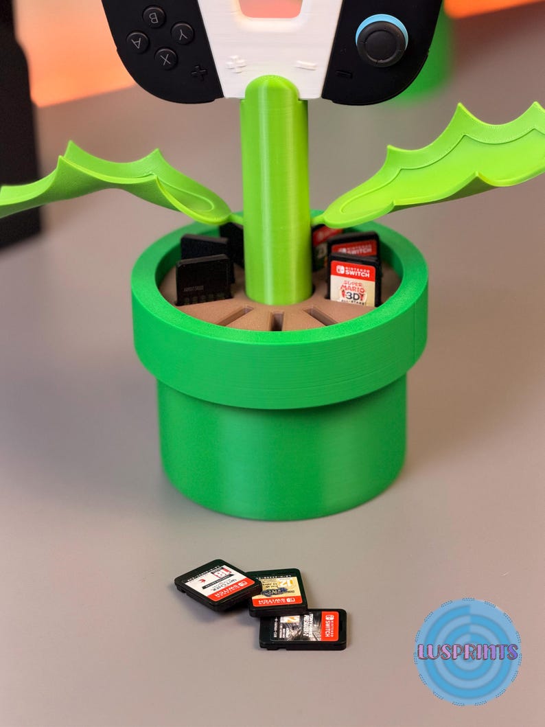 May include: A green and brown 3D-printed video game cartridge holder shaped like a flower pot, with a Nintendo Switch controller on top. Several game cartridges are in the pot, and three are in front of the pot.