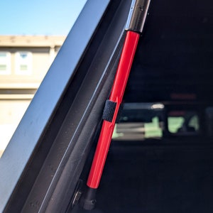 May include: A close-up of a black vehicle component featuring a vibrant red support strut. The strut is cylindrical with a black end and a black velcro strap. The vehicle's interior is dark, with the sky visible in the background.