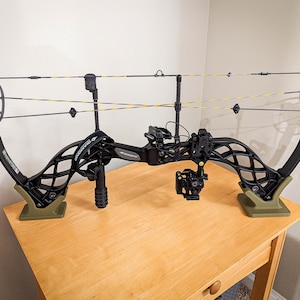 May include: A black compound bow with yellow and black strings, resting on a green stand. The bow features a sight, stabilizer, and a grip. The bow is on a wooden table.