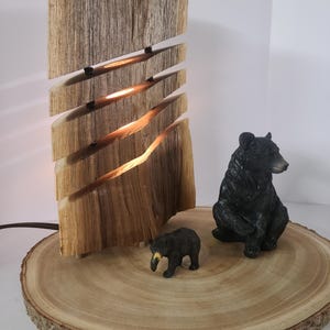 Può includere: Una lampada rustica in legno dal design unico, con intagli orizzontali che emettono luce calda. Due piccole statuette di orsi neri sono posizionate su una base a fetta di legno, creando un tema ispirato alla natura.