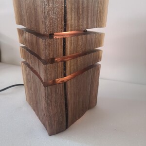 Può includere: Una lampada in legno fatta a mano con un design unico a strati. La lampada è realizzata in legno naturale con venature visibili e accenti scuri. La luce calda brilla attraverso le aperture orizzontali, creando un'atmosfera accogliente. La lampada è collegata con un cavo nero.