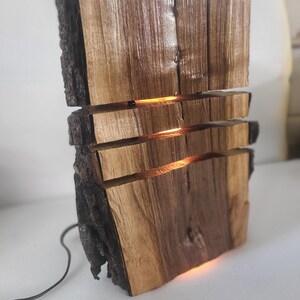 Può includere: Una lampada in legno fatta a mano con bordo in corteccia naturale. La lampada è composta da diversi pezzi di legno impilati con fessure orizzontali illuminate. La luce calda brilla attraverso le aperture, creando un'atmosfera accogliente.