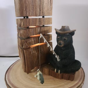 Può includere: Lampada rustica in legno con una statuina di orso nero con cappello da cowboy che pesca. La lampada ha un design in legno impilato con illuminazione calda. La base è una fetta di legno.