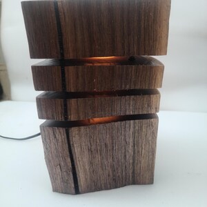 Può includere: Lampada da tavolo in legno fatta a mano con un bagliore caldo e invitante. La lampada è costruita con blocchi impilati di legno marrone scuro, con una linea nera. La luce emana dagli spazi tra i blocchi, creando un'estetica rustica unica.