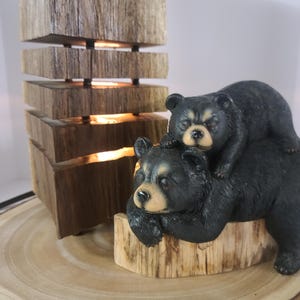 Può includere: Una lampada rustica in legno con un design impilato, che emette luce calda. Una statuina di orso nero con un cucciolo sulla schiena poggia su una base di legno, aggiungendo un tocco di ispirazione naturale.