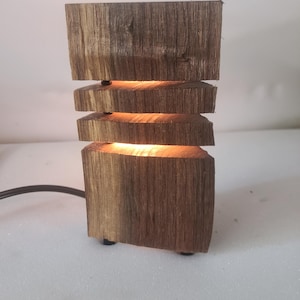 Può includere: Lampada in legno fatta a mano con un design unico. La lampada è costruita con pezzi di legno grezzi e impilati, con una luce calda che emana dagli spazi tra di essi. La lampada ha una base rettangolare e un cavo di alimentazione scuro. Altezza: 20 cm.