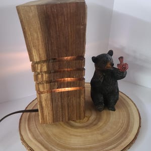 Può includere: Una lampada rustica in legno con un design intagliato, che emette un bagliore caldo. Una piccola statuina di orso nero che tiene un uccello rosso si trova accanto alla lampada su una base a fetta di legno. La lampada ha un cavo di alimentazione marrone.