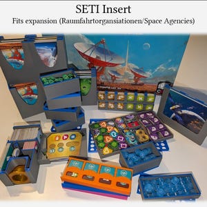 Pode incluir: Um inserto SETI cinza e azul para expansão de jogo de tabuleiro. O inserto apresenta vários compartimentos e bandejas para organizar os componentes do jogo, com um cenário temático espacial. O texto "SETI Insert" é visível.
