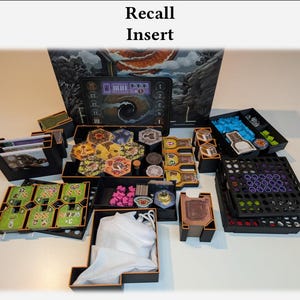 Op de afbeelding: Een verzameling zwarte spelcomponentenorganizers, gevuld met verschillende spelstukken, kaarten en tokens. De spelstukken omvatten zeshoekige tegels, kleine roze stukjes en andere kleurrijke spelcomponenten. De tekst "Recall Insert" is bovenaan zichtbaar.