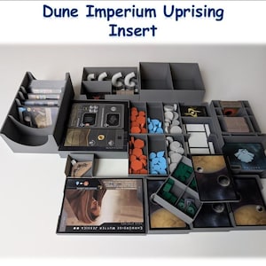 Puede incluir: Un inserto gris para el juego de mesa Dune Imperium Uprising. El inserto presenta varios compartimentos para cartas, fichas y componentes del juego. La imagen incluye el texto "Dune Imperium Uprising Insert".