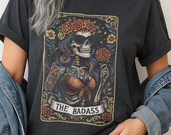 May include: Dark gray t-shirt featuring a tarot card-style graphic of a skeleton with a floral crown and sunglasses. The graphic includes roses, flowers, and the text "THE BADASS". The shirt has rolled-up sleeves.