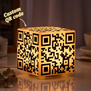 Puede incluir: Una lámpara de forma cúbica blanca con un diseño de código QR en blanco y negro. La lámpara está iluminada y proyecta un brillo cálido.