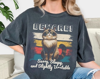 May include: A dark gray t-shirt with a vintage-style graphic of a cat with the text "BEWARE! Sassy, Sleepy, and Slightly Unstable".