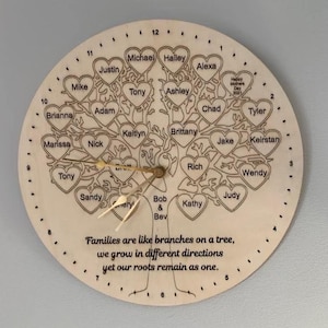 May include: A wooden clock with a tree design, featuring heart-shaped leaves with names. The clock hands are gold-coloured. The text "Families are like branches on a tree..." is at the bottom. The clock is approximately 30 cm in diameter.