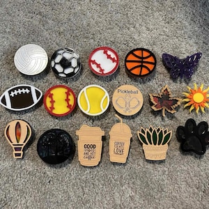 May include: A selection of decorative charms in diverse shapes, such as a volleyball, football, baseball, basketball, butterfly, American football, baseball, tennis ball, pickleball, leaf, sun, hot air balloon, black circle, coffee cup with text, coffee cup with text, succulent, and paw print.