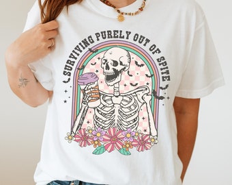 May include: White t-shirt with a graphic of a skeleton holding a coffee cup and the text "surviving purely out of spite". The graphic is surrounded by a rainbow arch and flowers.