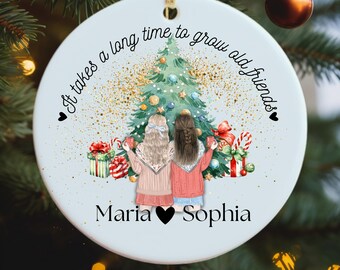 May include: A white ceramic Christmas ornament with a gold string for hanging. The ornament features a festive illustration of two friends holding mugs in front of a Christmas tree. Text reads "It takes a long time to grow old friends" and names "Maria" and "Sophia".