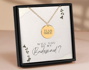 May include: A gold necklace with a round pendant that says "Team Bride" in a black box with a white background. The box also has the text "Will you be my Bridesmaid?" in black cursive writing.