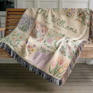 May include: A cream-coloured throw blanket with a floral design and the text "The Williams Est. 2024" in green. The blanket features a grid of floral illustrations, including daisies, tulips, and lavender, with a fringed edge.