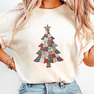 May include: An ivory short-sleeved t-shirt featuring a central graphic of a Christmas tree composed of various patchwork fabric patterns in red, green, blue, and white, topped with a star. The word Ivory is printed in the bottom corner.