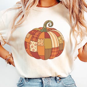 Può includere: Una t-shirt a maniche corte color avorio con una grande grafica di una zucca composta da vari motivi patchwork in tonalità arancione, marrone e verde tenue. Il design della zucca include sezioni floreali, a quadri e a righe con un picciolo.