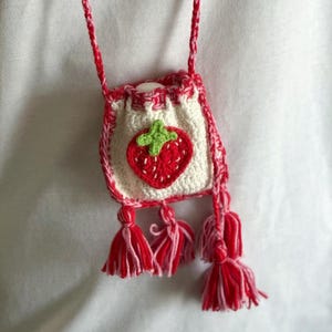 May include: A handmade crochet strawberry bag with a red and white strap. The small bag features a red strawberry applique with a green stem on a white background. Red and white tassels hang from the bottom.