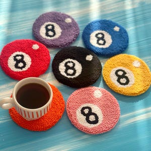 May include: A set of colourful, round coasters, each with a number 8 billiard ball design. The coasters are in shades of red, purple, blue, black, yellow, pink, and orange. A white teacup with dark liquid rests on an orange coaster.